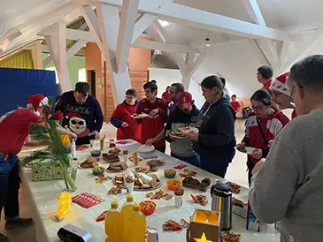 Eine Gruppe von Menschen feiert Weihnachten an einem langen Tisch voller weihnachtlichen Snacks und Getränke. Viele tragen rote Kleidung, einige Weihnachtsmützen.