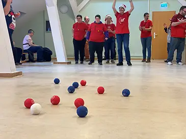 Eine Gruppe von Menschen spielt Boccia in der Remise. Einige stehen bereit, andere sitzen am Rand und beobachten das Spiel.